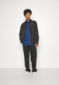 Schwarze leichte Jacke über einem blauen Hemd mit Knöpfen, kombiniert mit schwarzen Hosen und weißen Sneakers. Das Model steht vor einem einfarbigen Hintergrund.