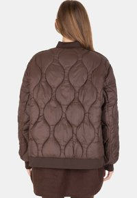 Donna che indossa una giacca trapuntata marrone con un motivo a foglia, vista di spalle, con i lunghi capelli che cadono sul colletto.