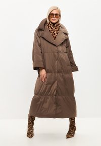 Cappotto puffer oversize marrone con un ampio colletto, caratterizzato da un design a strati e tasche a filetto, abbinato a una sciarpa con stampa leopardata e stivali.