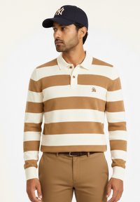 Suéter a rayas en beige y marrón claro con cuello, que presenta un logotipo en el pecho. Combinado con pantalones color cuero y una gorra azul marino.