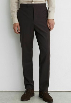 Mann trägt maßgeschneiderte dunkelbraune Hose, beiges Hemd und dunkelbraune geschnürte Lederschuhe und steht vor einer weißen Wand.