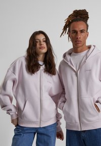Felpa rosa con zip realizzata in morbido tessuto, con tasche frontali e polsini a coste. Il nome del marchio è ricamato in una tonalità sottile.