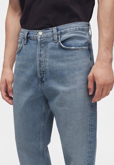 Homme portant un jean en denim bleu clair et une chemise noire, debout les mains détendues le long du corps.