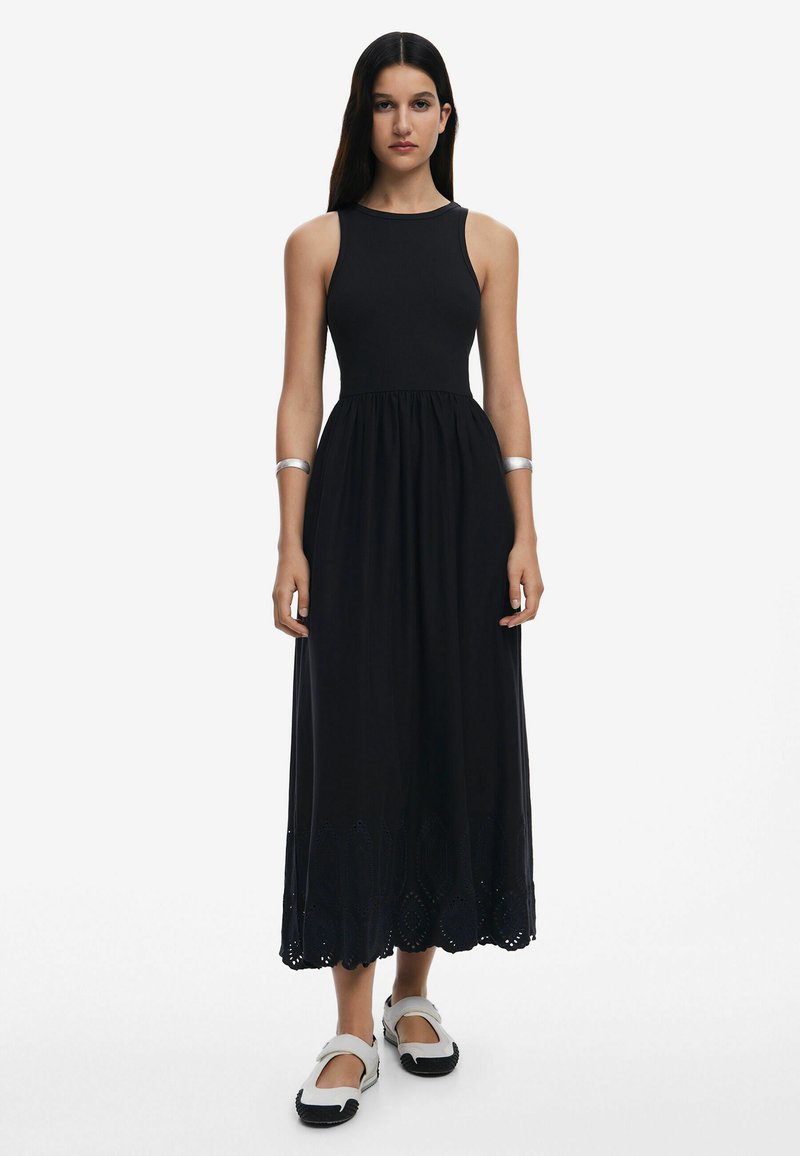 Vestido maxi sin mangas negro con un cuerpo ajustado y una falda en línea A que presenta intrincados diseños recortados a lo largo del dobladillo. Combinado con sandalias blancas.