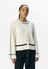 Cardigan bianco a coste con accenti neri, scollo rotondo e bottoni perlati. Maniche lunghe con finitura testurizzata. Silhouette aderente.