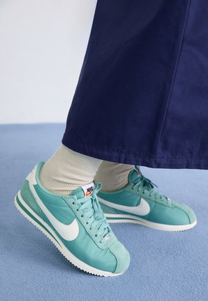 Voeten met lichtblauwe Nike-sneakers met witte swoosh, beige sokken en donkerblauwe broek, staand op een lichtblauw tapijt.