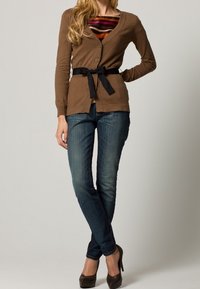 Brauner Cardigan mit Knöpfen und einem schwarzen Bindegürtel, getragen über einem gestreiften Oberteil und kombiniert mit dunkelblauen Jeans und braunen Absätzen. Glatte Textur.