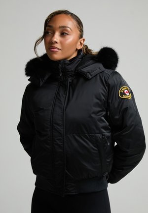 Femme en veste d'hiver noire avec capuche bordée de fourrure et écusson Canada sur la manche, regardant légèrement de côté sur fond uni.