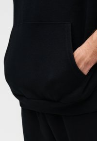 Χέρι στην τσέπη μαύρου φούτερ με κουκούλα, με εμφανή μπροστινή τσέπη και μαύρο παντελόνι, σε απλό φόντο.