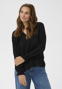 Femme aux longs cheveux bruns portant un haut à manches longues noir et un jean bleu, debout avec les bras croisés sur fond blanc.