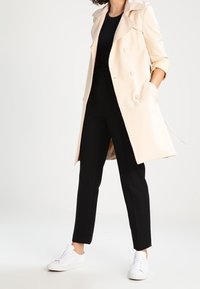 Beige doppelreihiger Trenchcoat mit Reverskragen, getragen über einem schwarzen Oberteil und schwarzen Hosen. Kombiniert mit weißen Sneakers.