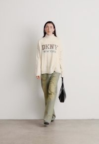 Maglione a maglia color crema con testo "DKNY NEW YORK", abbinato a jeans svasati verde chiaro e una borsa nera con finitura testurizzata.