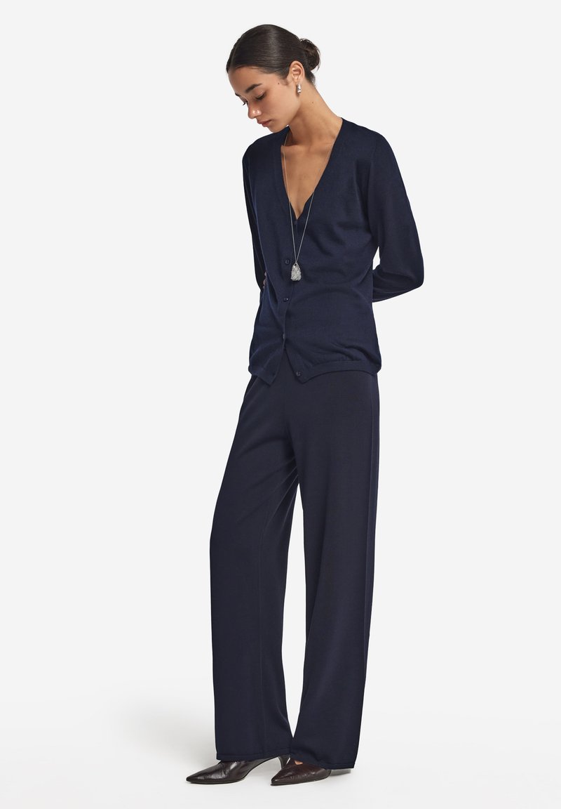 Marinblå cardigan med djup V-ringning, långa ärmar och knäppning, ihop med vida marinblå byxor och spetsiga mörka skor.