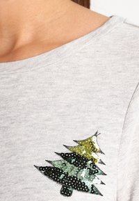 Sudadera gris con un diseño de árbol de Navidad de lentejuelas verdes y negras cosido en el pecho. La tela tiene un aspecto suave y texturizado.