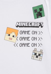 Biely tričko s pixelovanými postavami z Minecraftu: zelený creeper, oranžová postava a pes, s textom "HRA ZAČÍNA" v čiernej farbe.