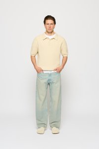 Cămașă polo tricotată ribbed de culoare crem, cu mâneci scurte, asortată cu blugi din denim ușori, de culoare albastru deschis, cu croi lejer. Încălțămintea de culoare crem completează ținuta.