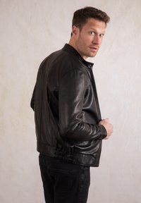 Black leather jacket with a zippered front, features ribbed cuffs and a gathered hem. Styled with black jeans and a neutral background.