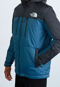 Osoba nosząca niebieską i czarną, ocieplaną kurtkę zapinaną na zamek z logo The North Face na piersi, z jedną ręką w bocznej kieszeni.
