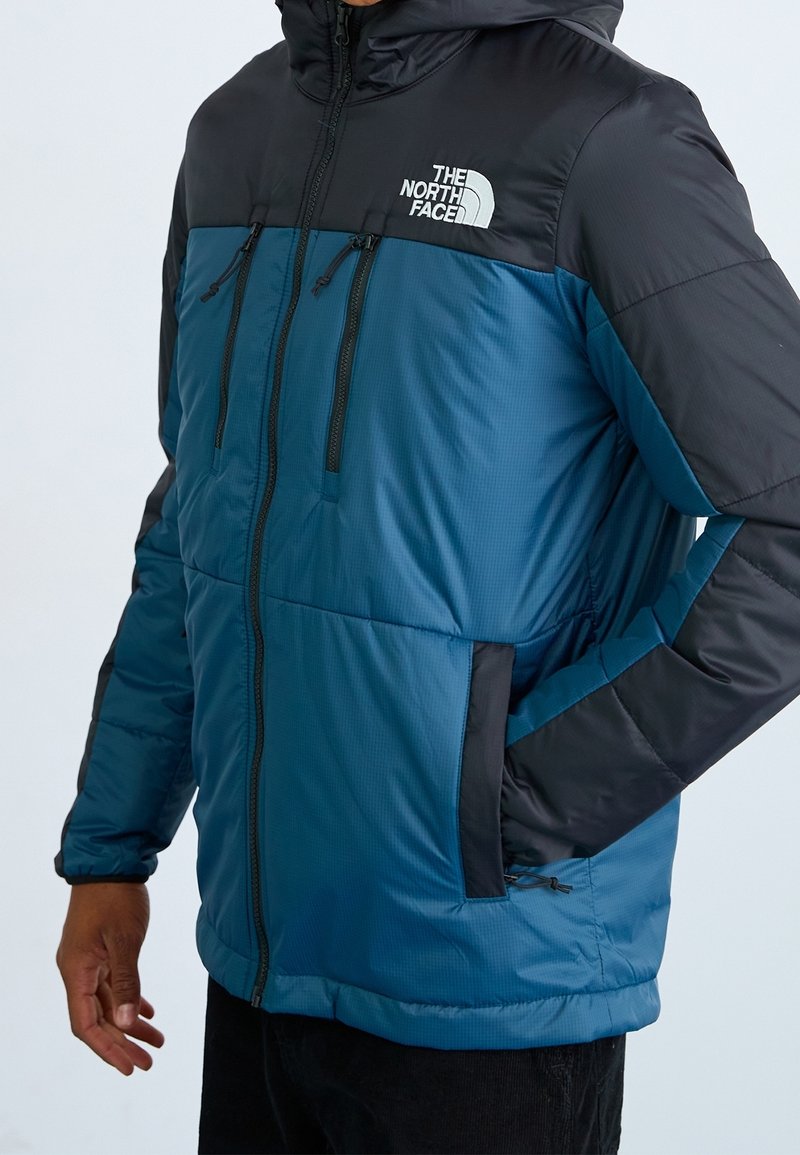 Osoba nosząca niebieską i czarną, ocieplaną kurtkę zapinaną na zamek z logo The North Face na piersi, z jedną ręką w bocznej kieszeni.
