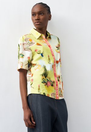 Woman with braided hair wears short-sleeve button-up shirt with colorful bird, cactus, lipstick, and desert-themed prints, paired with dark pants.