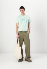 Camiseta verde claro con un fuerte logo de "GUESS", combinada con pantalones olivo de corte suelto. Un bolso de tela color crema y zapatos marrones completan el look.