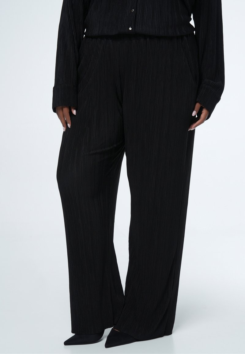Femme portant un pantalon large plissé noir, des chaussures noires à bout pointu et un haut noir assorti à manches longues avec des boutons, debout sur un sol blanc.
