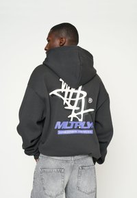Zwarte hoodie met een grote witte grafische afbeelding en paarse tekst op de achterkant. Heeft een losse pasvorm en een capuchon met trekkoord. Draagt met grijze jeans.