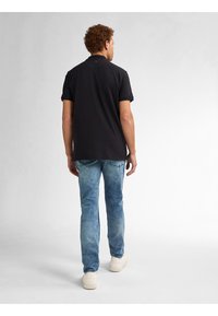 Camiseta polo negra de manga corta, combinada con jeans deslavados de color azul claro y zapatillas blancas, vista desde la parte posterior sobre un fondo neutro.