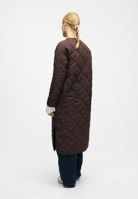 Femme avec une tresse blonde porte un long manteau matelassé marron foncé, des manches en tricot blanches, un pantalon bleu foncé et des chaussures beiges, se tenant de dos.