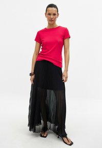 T-shirt rosso aderente abbinato a una lunga gonna plissettata in chiffon nera. La gonna ha una texture trasparente e un design fluido. La modella indossa sandali neri con suola piatta.