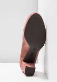 Une chaussure à talon pour femme en daim rose reposant sur le côté, montrant la semelle noire avec le nom de la marque "Unisa" visible.