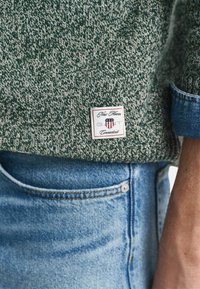 Grüner und weißer gestrickter Pullover mit sichtbarem Markenlabel. Kombiniert mit blauen Jeans, die ein Taschendetail aufweisen.
