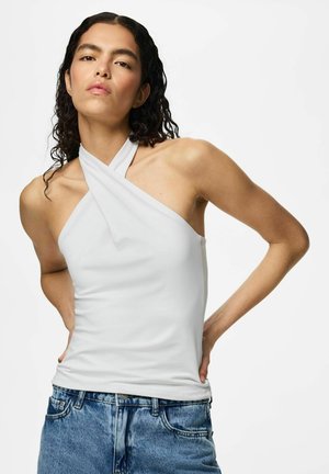 Femme aux cheveux bouclés et foncés, portant un haut blanc sans manches à col halter et un jean bleu, debout avec les mains dans le dos devant un fond uni.