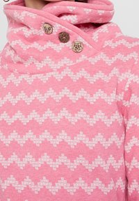 Felpa rosa con colletto alto, caratterizzata da un motivo a zigzag bianco. Tre bottoni decorativi arricchiscono il design su un lato.