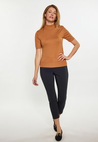 Ruskea lyhythihainen neule, jossa on korkea kaulus ja nappidetalji, yhdistettynä mustiin slim fit -housuihin ja mustiin tasapohjaisiin kenkiin, joissa on metalliyksityiskohtia.