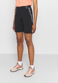 Osoba nosící černé krátké kalhoty po kolena s bočním zipem, bílé kotníkové ponožky a růžové sportovní boty, stojící na šedé podlaze.