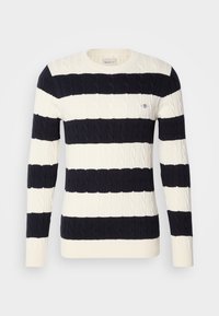 Tamsiai mėlynas ir kremowy dryžuotas megztinis su kabelių mezgimo raštu, grioveliais apdailos iškirptėje, rankogaliuose ir apačioje, su maža logotipo ženkleliu ant krūtinės.