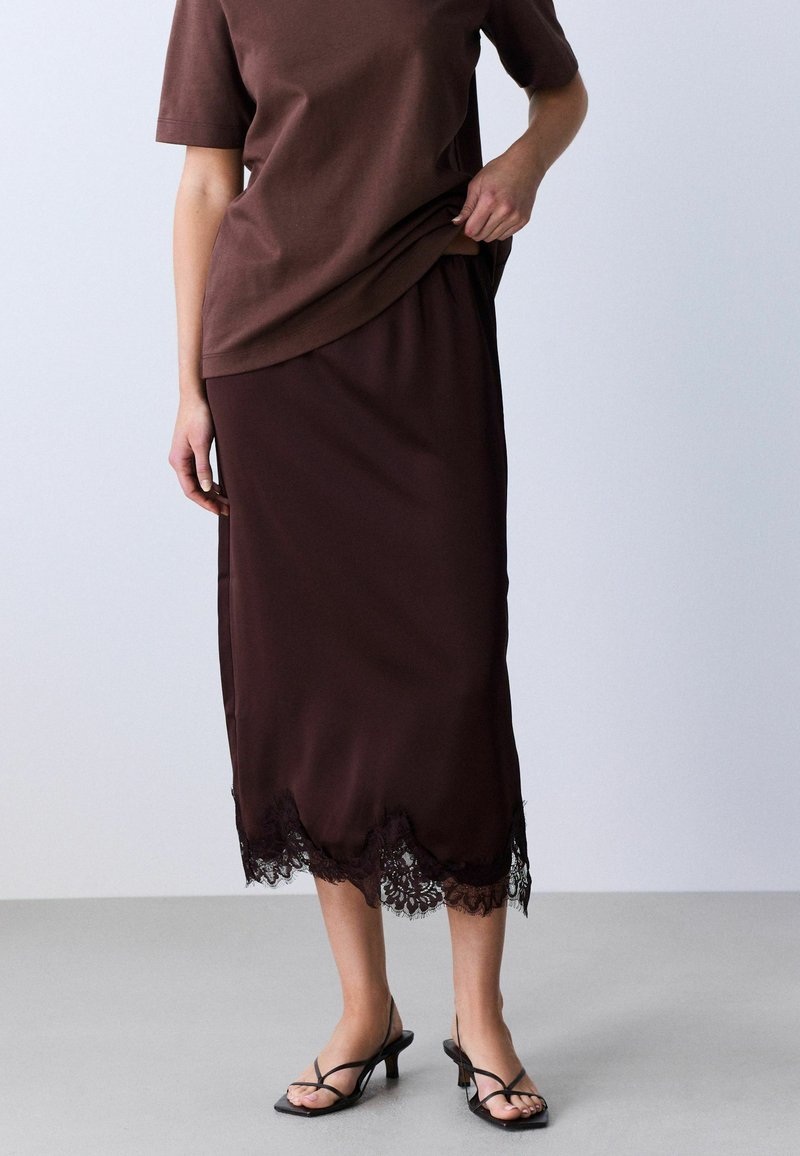 Femme portant une jupe midi marron foncé avec un ourlet en dentelle et un top à manches courtes assorti, associé à des sandales à talons noires à brides.