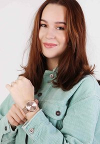 Roségoldene Uhr mit rundem Ziffernblatt und Metalldrahtarmband, am Handgelenk getragen. Der Träger trägt eine hellgrüne Cordjacke mit Druckknöpfen.