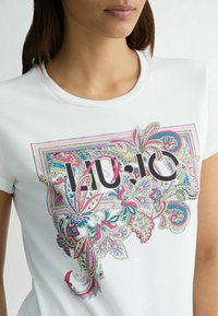 T-shirt bianco in cotone con una stampa paisley multicolore vibrante e testo nero audace, dotato di scollo rotondo e maniche corte.