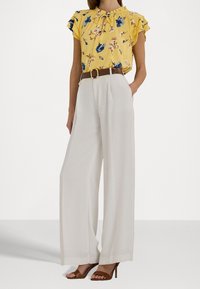 Gul blommig blus med volangärmar, kombinerad med högmidjade vita vida byxor och bruna klacksandaler.