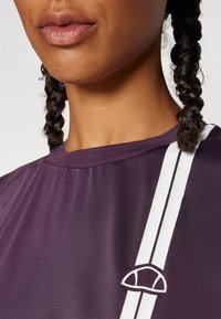 Μωβ αθλητική μπλούζα με στρογγυλή λαιμόκοψη, άσπρες λωρίδες στα πλάγια και ένα μικρό λογότυπο σε άσπρο κοντά στη λαιμόκοψη, φτιαγμένη από μαλακό, υφασματοποιημένο ύφασμα.