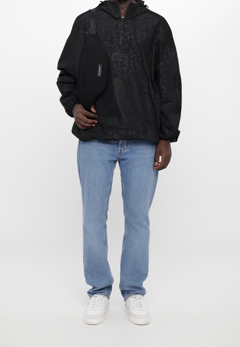 Man i svart mönstrad jacka, svart crossbody-väska, ljusblå jeans och vita sneakers som står mot en vit bakgrund.