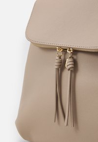 Zaino in finta pelle beige con chiusura superiore a zip, dotato di due frange decorative in pelle e dettagli in metallo dorato. Texture liscia.