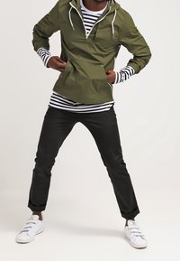 Osoba ubrana w oliwkową kurtkę z kapturem na czarno-białą prążkowaną koszulę, czarne spodnie i białe trampki, stojąca w rozkroku.