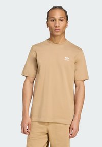 Tan kleur katoen t-shirt met een ronde hals, korte mouwen en een klein wit Adidas-logo op de linkerborst. Gladde textuur, relaxte pasvorm.