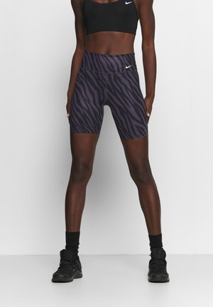 Sportowiec ubrany w czarny sportowy biustonosz i fioletowe szorty rowerowe z czarnym nadrukiem w zebrę o wysokim stanie, czarne buty sportowe i skarpetki.