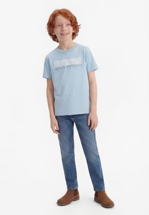 Rødthåret dreng iført en lyseblå Abercrombie and Fitch t-shirt, blå jeans og brune støvler, stående og smilende let til siden.