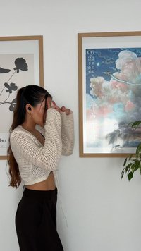 Cardigan de crema de ganchillo con un corte holgado y mangas anchas, combinado con pantalones negros de tiro alto. Arte mural y una planta en el fondo.