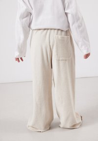 Enfant portant un pantalon ample en éponge de couleur crème avec une poche arrière et une chemise blanche à manches longues, debout sur un sol clair.